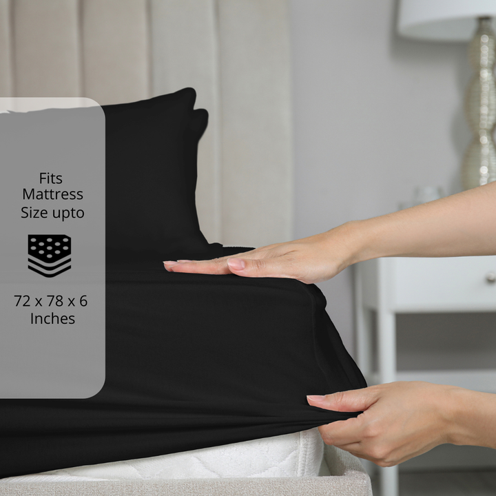 Black fitted bedsheet being stretched over mattress corner with size label showing fits mattress size up to 72 x 78 x 6 inches.  