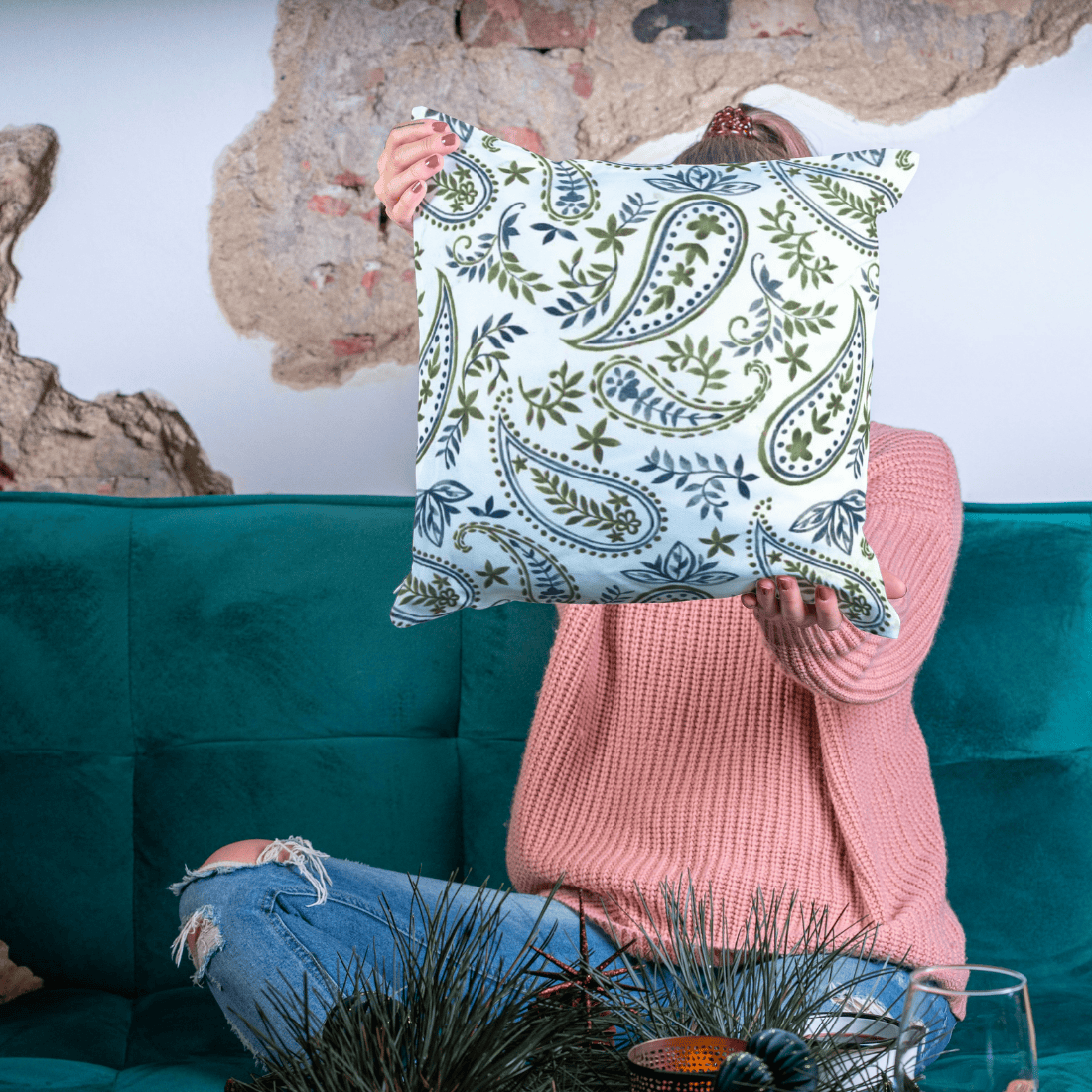 Person holding a decorative pillow with a paisley pattern in front of a teal couch.