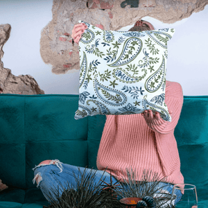 Person holding a decorative pillow with a paisley pattern in front of a teal couch.