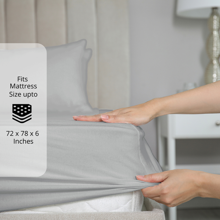 Silver fitted bedsheet being stretched over mattress corner with size label showing fits mattress size up to 72 x 78 x 6 inches.  
